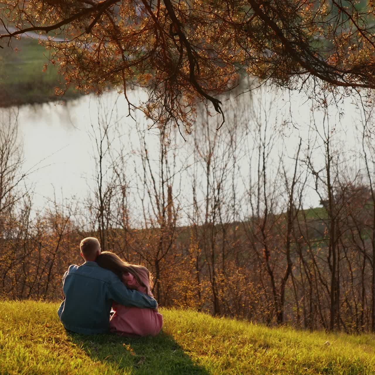 Couple in love resting among autumn nature. Young man and woman sitting on the hill and admiring the river at sunset