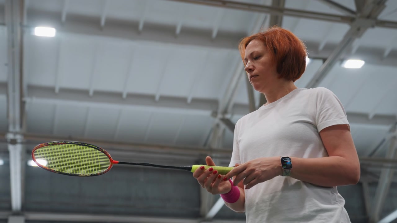 Redhaired white woman inspecting racket during indoor badminton warmup, coach inspecting grip and strings, focused expression, gymnasium lighting, slow deliberate movements, quiet preparation