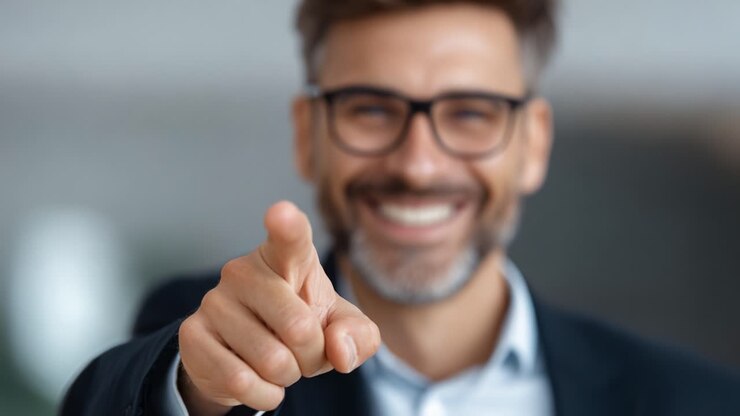 Uomo sicuro di sé che sorride e indica direttamente la fotocamera, trasmettendo carisma ed entusiasmo in un ambiente professionale