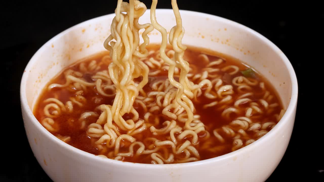 Close-up of fork lifting steaming instant noodles from bowl of soup on black background