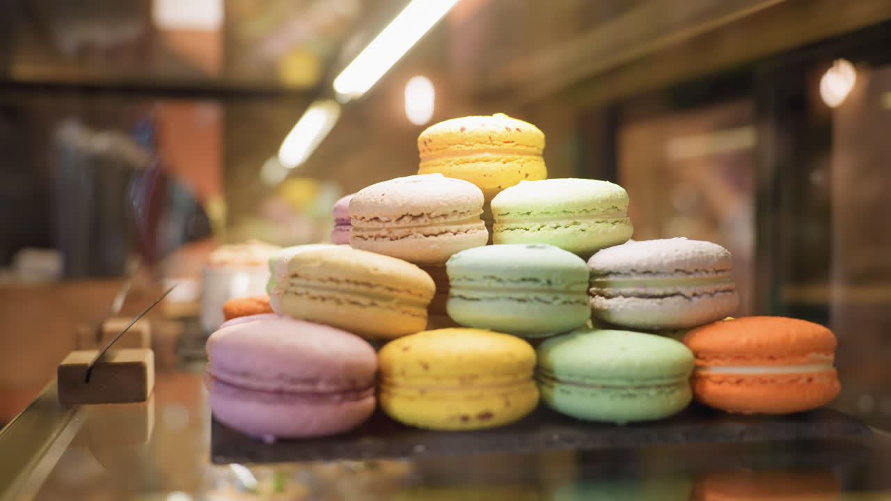 close up display of colorful assorted macarons arranged in pyramid on black slate in dessert showcase under warm lighting, soft reflections on glass create inviting bakery