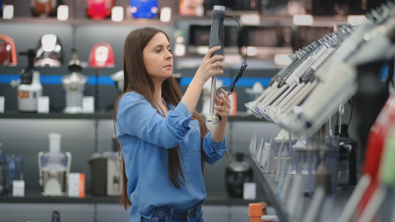 en una tienda de electrodomésticos, una mujer morena con una camisa azul toma una licuadora en sus manos y considera el diseño y las especificaciones.