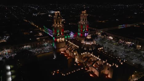morelia cathedral at night, lit with the patriotic colors green, white, and red. drone shot during the month of september.