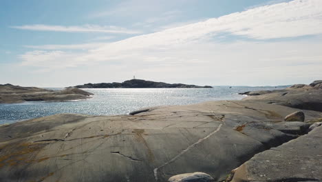 Scenic-crisp-summertime-view-of-shimmering-and-shiny-ocean-sea-water-from-flat-smooth-shoreline-rock-on-bright-blue-high-sunny-sky-day,-Bohuslan,-Sweden,-handheld-pan