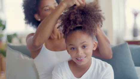 african american mother combing daughter's hair while little girl smiling and looking at the camera