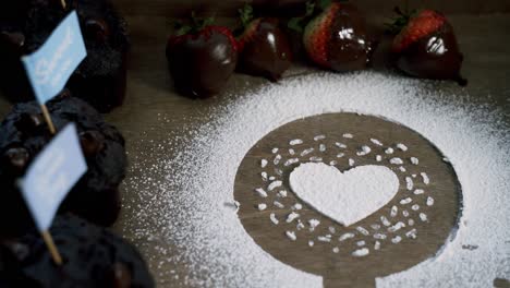 making a heart out of white confectionate powdered sugar with chocolate covered strawberries and muffins on the side