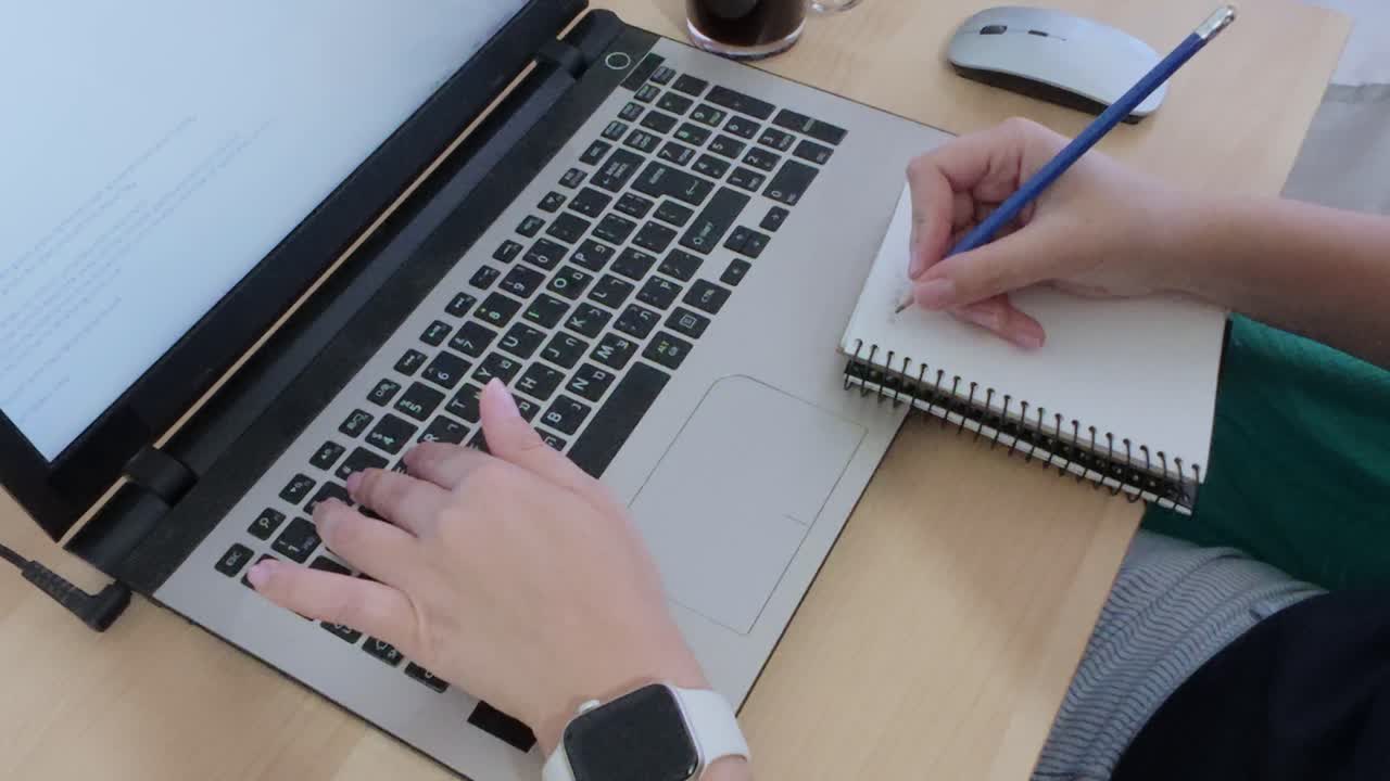 Close-up of hands multitasking, writing in a notebook while using a laptop with Hebrew text.