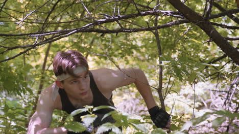 young man bending forward while walking under low hanging branch, elbow pushing aside thin wood limb, muscles tensing as he navigates dense forest undergrowth with focused determined expression