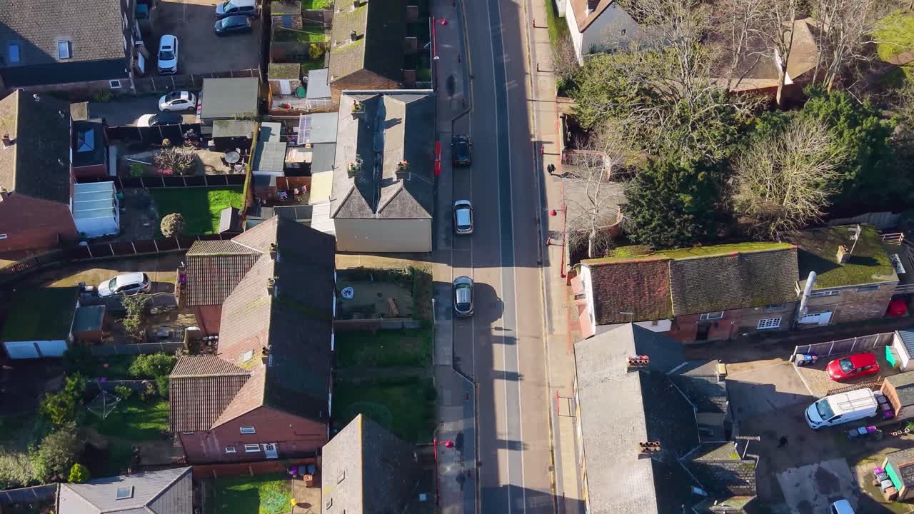Sorvolo con drone della via principale di Stanstead Abbots verso la stazione ferroviaria, che mostra le ombre dei negozi, traffico leggero, auto parcheggiate e segnaletica orizzontale in una piccola città inglese.