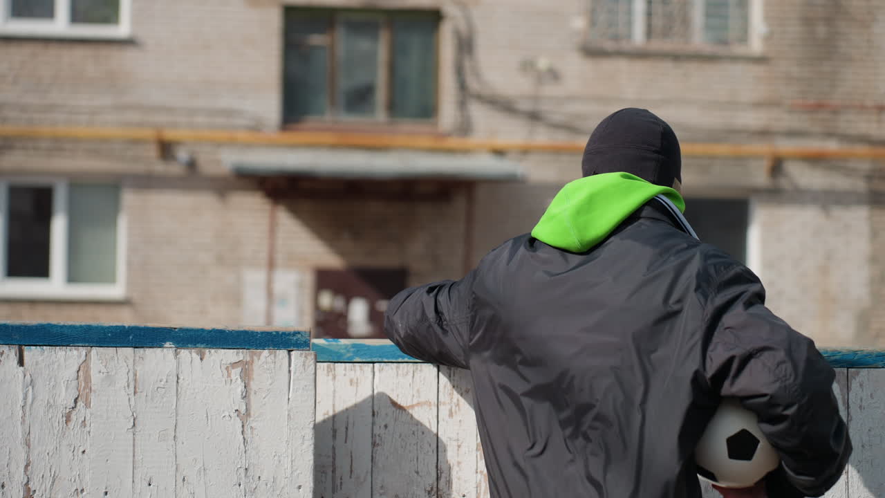 Un mentor adolescente blanco y un compañero de equipo en una cancha ruinosa, señalando y demostrando la técnica por encima de una barrera blanca, una pequeña pelota de fútbol, expresión atenta, momento de entrenamiento urbano, orientación de apoyo