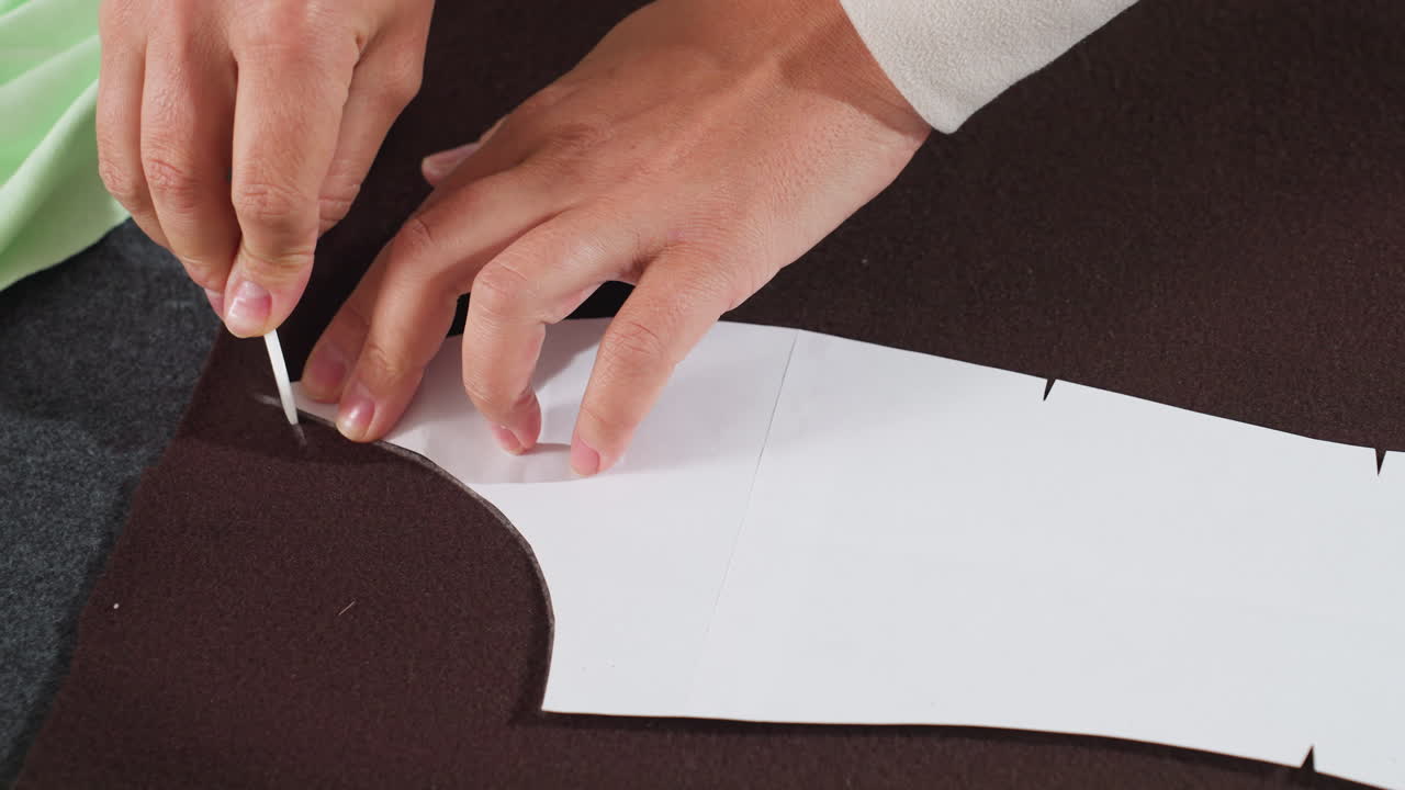 Fashion designer carefully uses tailors chalk to trace white paper pattern on dark brown fabric connecting lines precisely during dressmaking process inside bright creative sewing workspace