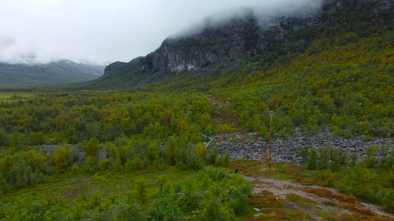 Aerial drone footage of rocky and forest covered Sweden landscape with a mountain in clouds in the background while a person is walking on rocky terrain in the foreground. Ground has dirt and is brown