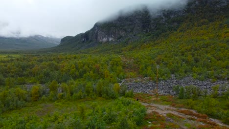 aerial drone footage of rocky and forest covered sweden landscape with a mountain in clouds in the background while a person is walking on rocky terrain in the foreground. ground has dirt and is brown