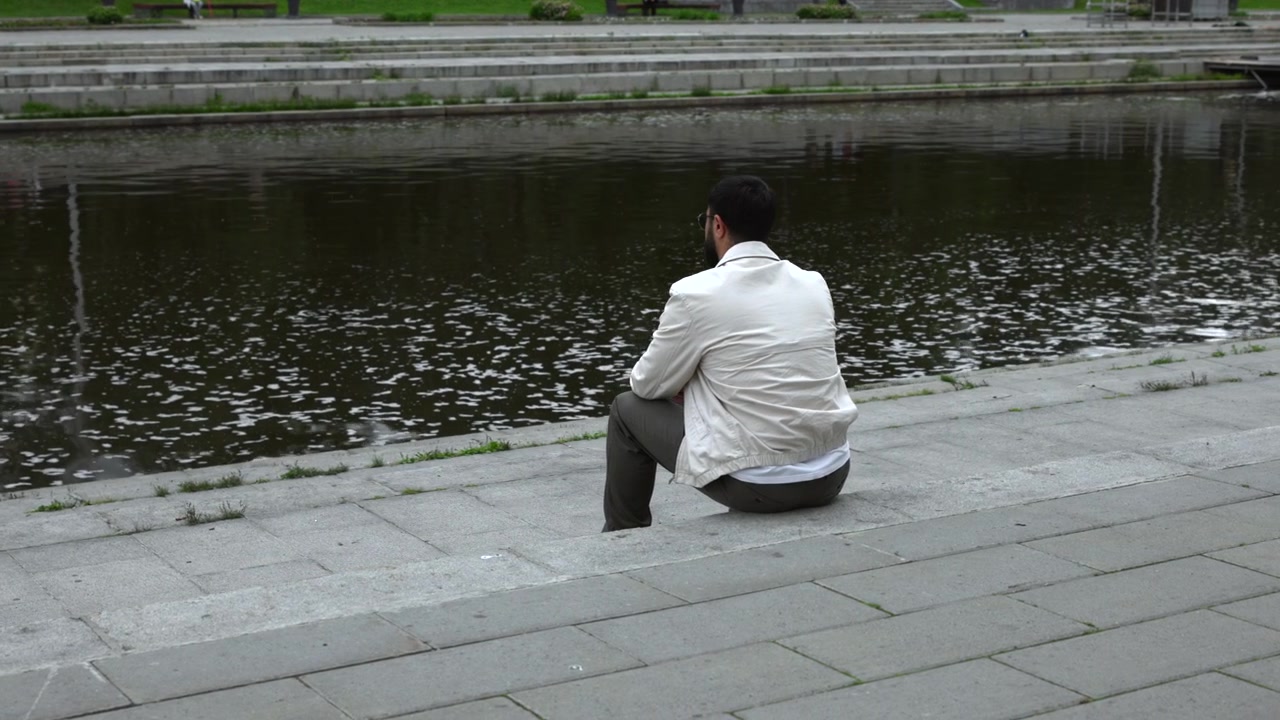 Homem adulto sentado em degraus de pedra ao lado de um lago escuro e reflexivo, inclinado ligeiramente para a frente com os braços apoiados nos joelhos, rodeado por texturas suaves de parque, água calma e luz do dia nublada.