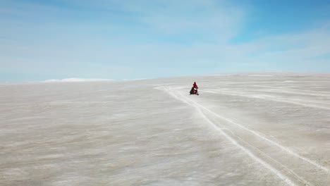 Aerial-view-of-a-person-doing-stunts-on-a-snowmobile,-on-the-white-surface-of-a-glacier-in-Iceland,-on-a-sunny-day