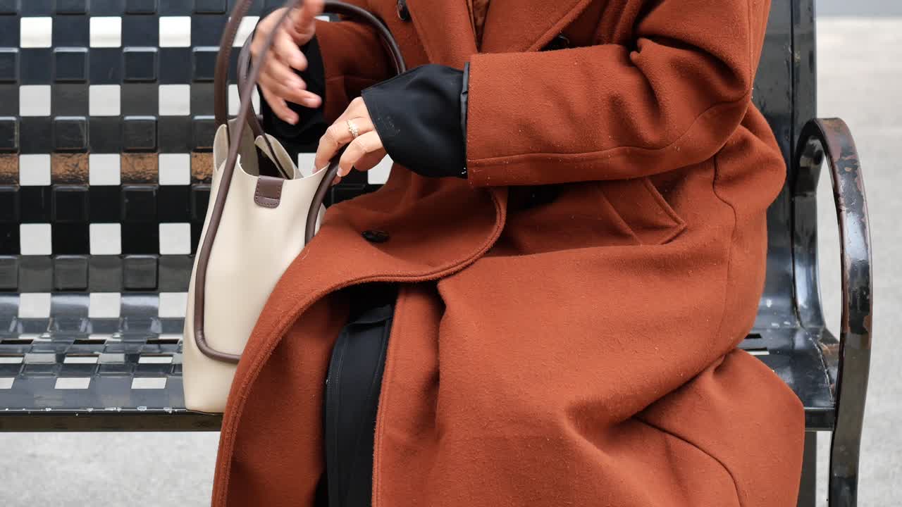 벤치에 앉아있는 여자, 드백에 손을 넣고, 갈색 코트를 입고