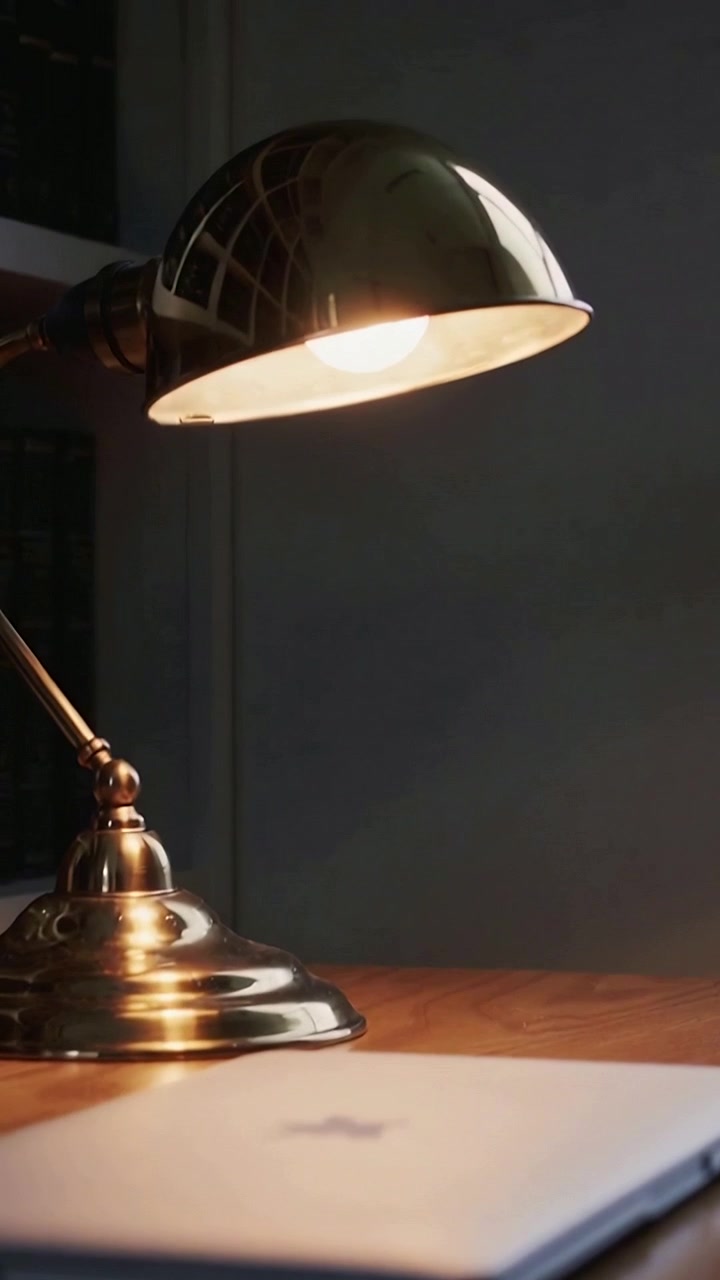 Video verticale: accensione di una lampada da scrivania in ottone che proietta una luce calda su un tavolo da studio, mostrando il logo del portatile