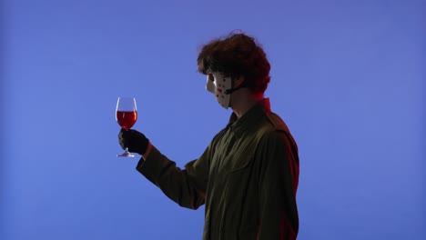 man in white mask holding a glass with red wine on blue background with red neon light image of