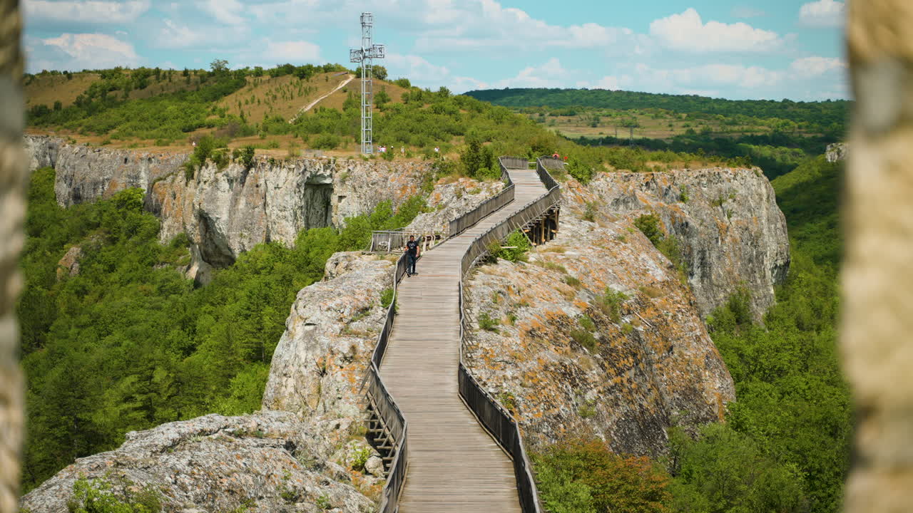 Man Walking On The Wooden Bridge With Orthodox Cross In The Distance Seen From Ovech Fortress In Bulgaria. - wide shot