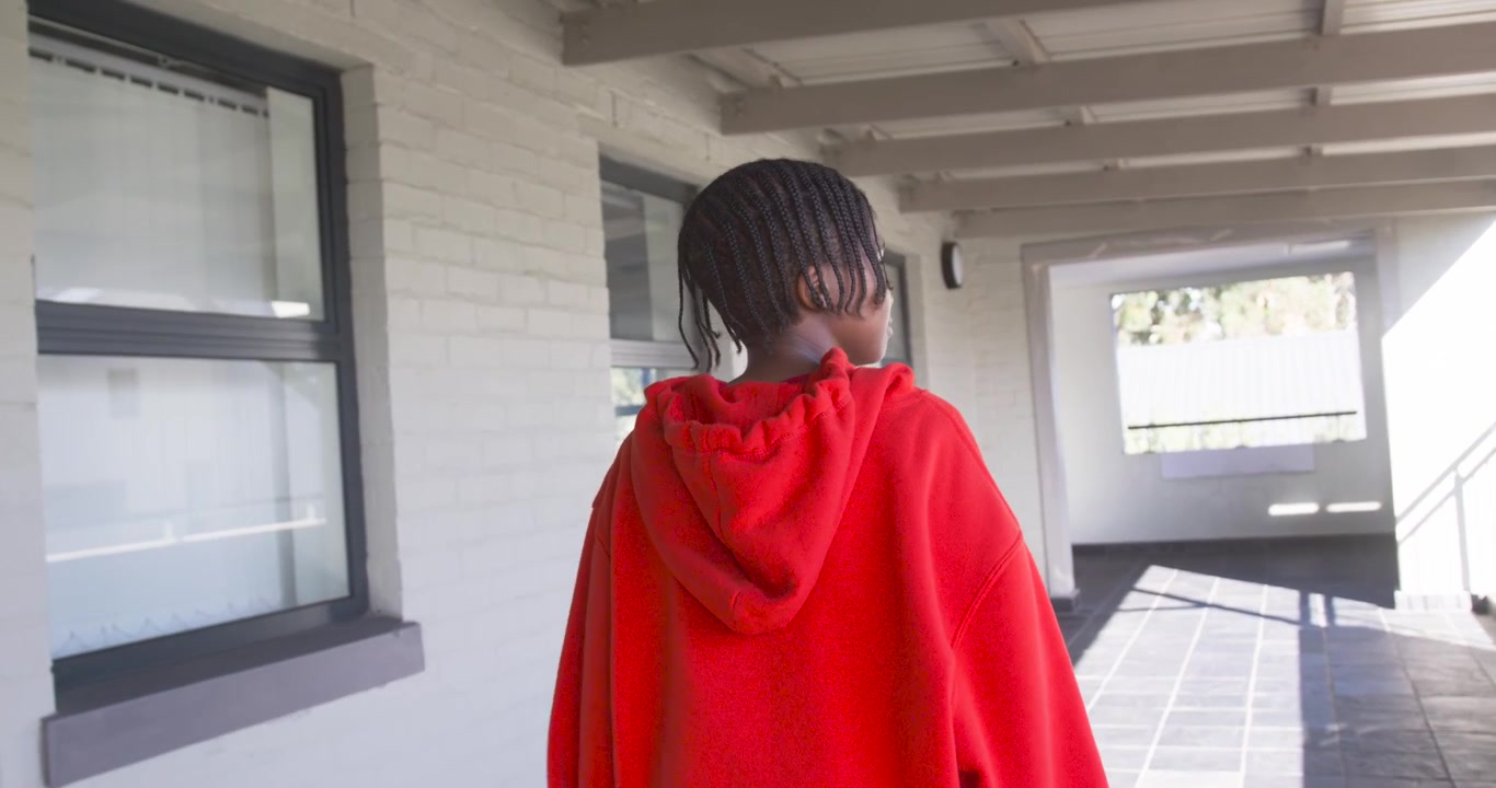 African teenage male walking down covered corridor wearing red hoodie, turning toward sunlit steps
