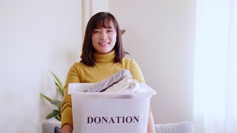 portrait of young asian woman looking at camera holding box with clothes for donation