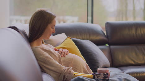 pregnant woman with prosthetic arm relaxing sitting on sofa at home with hot drink