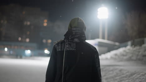 back view of individual dressed in winter attire walking across snow-covered surface at night, illuminated by strong stadium lights in urban environment, conveying solitude and cold atmosphere