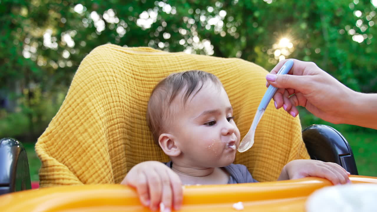 Beautiful hungry Caucasian toddler opens mouth willingly for a spoon with porridge. Mom's hand feeding her little son outdoors. Nature backdrop in blur.
