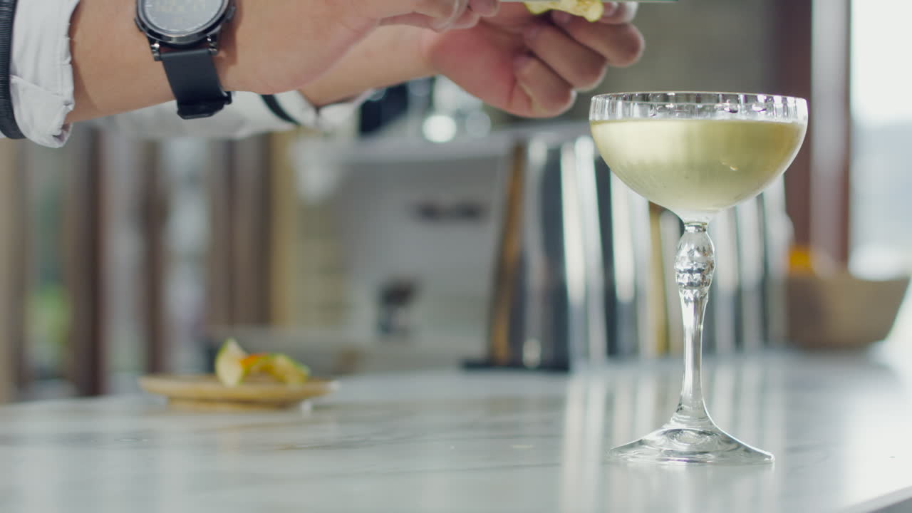 Close-up of a decorated cocktail glass next to an appetizer, with a bartender adjusting the items on the bar
