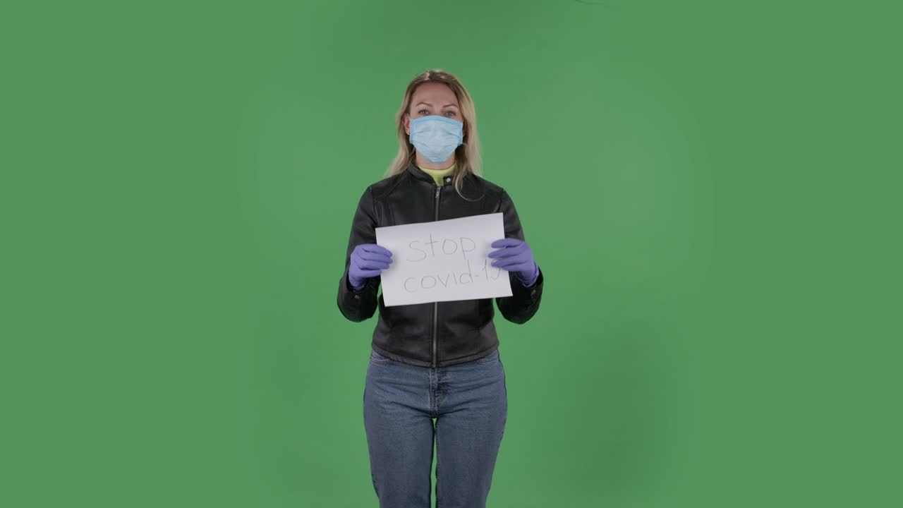 Portrait of beautiful young woman in medical mask is looking at camera and holding a poster Stop Kovid-19.Blonde with loose hair in a black jacket and jeans on a green screen in the studio. Health Protection Corona Virus Concept