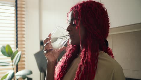 mujer afroamericana rizada y alegre bebiendo un vaso de agua dulce por la mañana