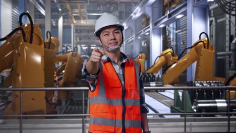 asian male engineer with safety helmet smiling and touching his chest then pointing at you while standing with industrial robotic arms working