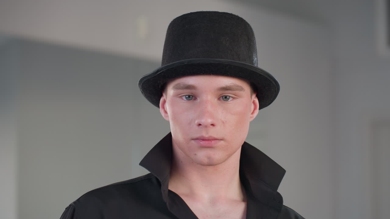 young man in black outfit and top hat stares confidently into camera inside loft studio with mirror reflections highlighting neck tattoo and popped collar exuding modern style and suave attitude