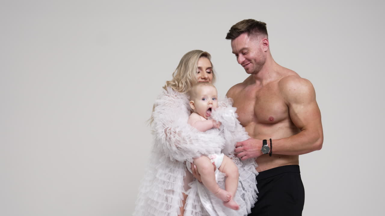 Happy smiling woman in white robe waves her cute little baby. Muscular man stands behind embracing his wife with child. White backdrop.