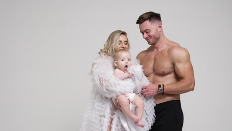 happy smiling woman in white robe waves her cute little baby. muscular man stands behind embracing his wife with child. white backdrop.