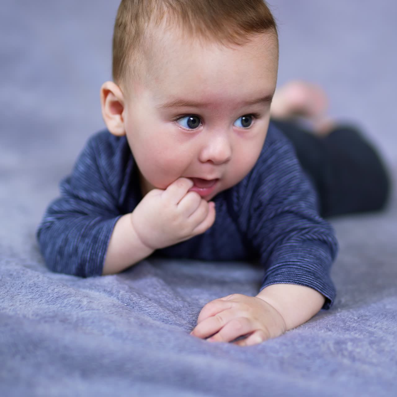 Adorable Caucasian child lies on belly with his finger in mouth. Lovely sweet baby resting peacefully on the grey plaid. Close up. Blurred backdrop