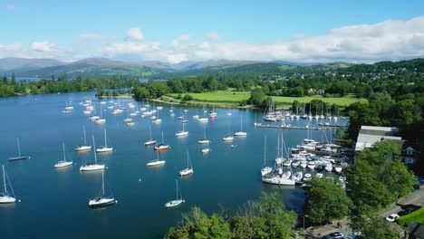 Absteigender-Drohnenblick-über-Bootsliegeplätze-Und-Yachten-Auf-Dem-Lake-Windermere-Am-Fährhafen-Mit-Bunten-Bäumen,-Bergen-Und-Blauem-Himmel-Mit-Weißen-Wolken-An-Einem-Sonnigen-Sommermorgen