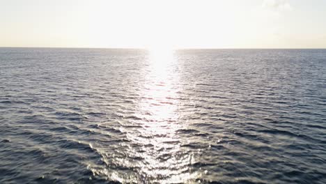 una carretilla aérea hacia el sol poniéndose como un destello de luz que se extiende reflejando a través de la superficie del agua del océano