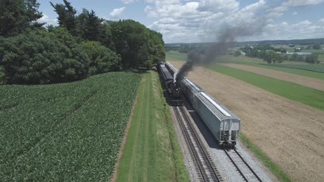 Vista-Aérea-De-Las-Tierras-De-Cultivo-Y-El-Campo-Con-Un-Tren-De-Vapor-Antiguo-Que-Se-Hincha-Para-Ponerse-En-Marcha-En-Un-Día-Soleado-De-Verano