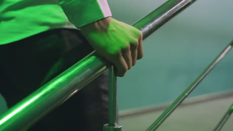 man face not visible, wearing white shirt and grey trousers, walking on stairs while gripping iron rail. green lighting casts modern, sleek ambiance, emphasizing movement and determination in an urban