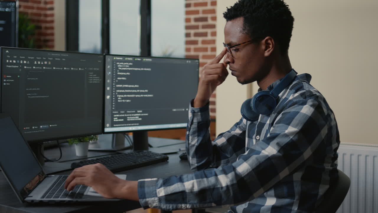 Programer thinking while touching beard and fixing glasses while typing on laptop sitting at desk