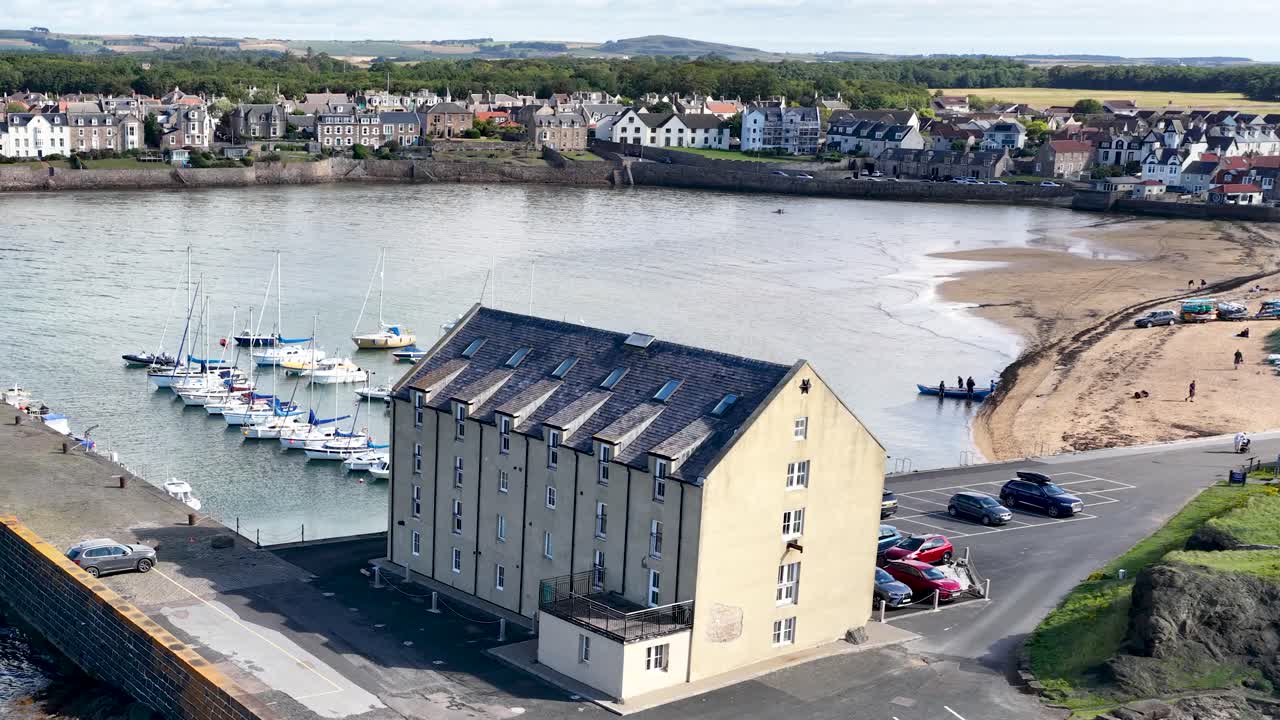 Drone pans across marina, old building, parked cars, and waterfront village in bright daylight