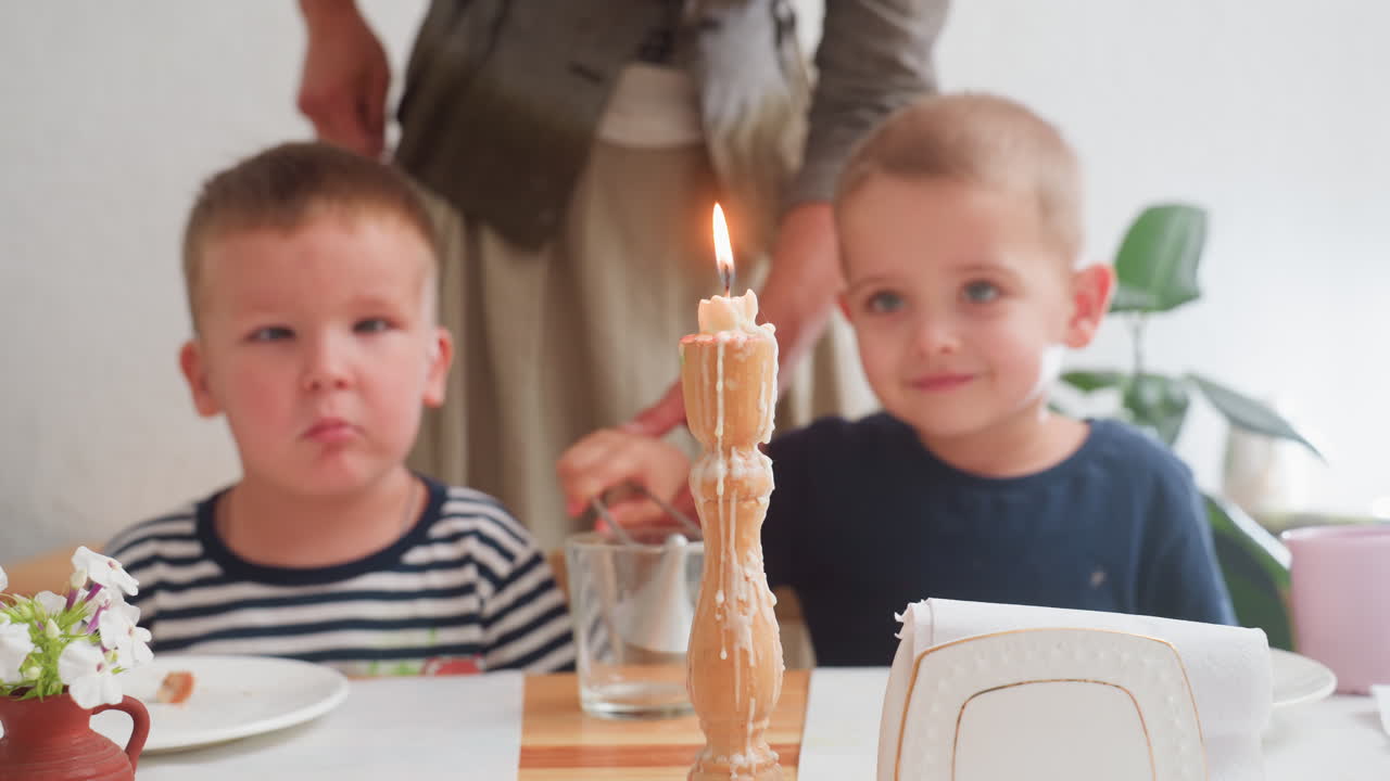 Educator lights candle using lighter as two young boys watch attentively during kindergarten lesson, one with striped shirt sitting beside classmate at wooden table surrounded by dishes