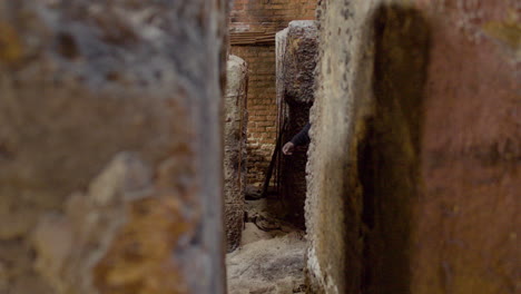 the camera focuses on a girl running away from a man in a balaclava and holding an ax behind the walls of a ruined building