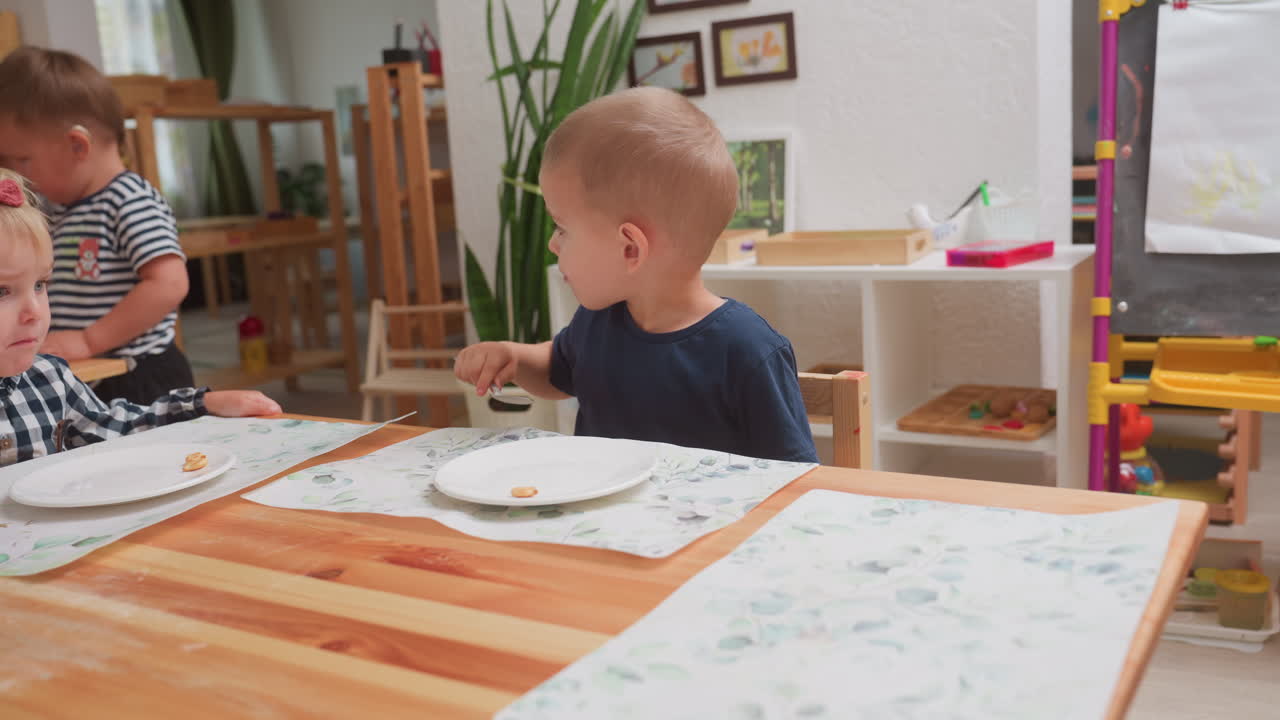 Bambino curioso con maglietta blu scuro tiene un cucchiaio in mano e guarda distratto da un'altra parte durante il pranzo con altri bambini in un ambiente interno luminoso, con piatti vuoti e tovagliette floreali su un tavolo di legno