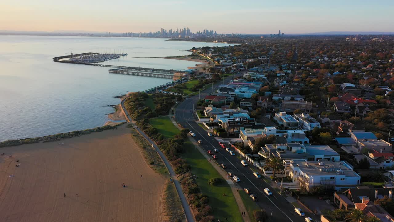 Drone video flying over Brighton's beach and road along the coast, with Melbourne's beautiful City skyline in the far horizon at sunset