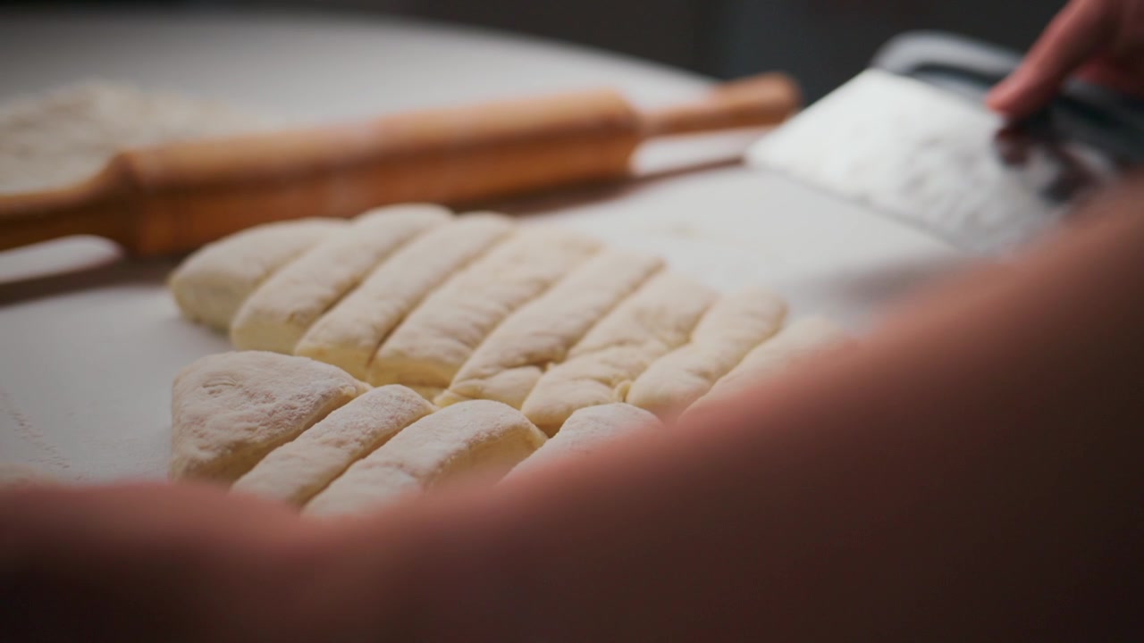 Close-up of hands slicing dough into pieces on a white surface. Rolling pin and knife are nearby, ready for use. Fingers press gently as dough separates cleanly. Scene conveys calm focus, precision