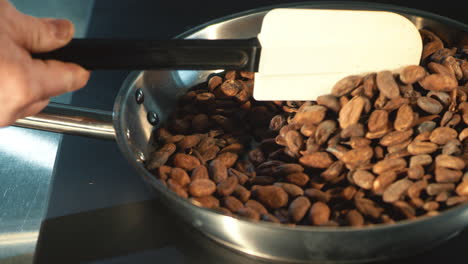 human hand stirring with spatula cocoa grains roasting on pan.