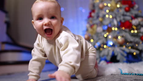 beautiful baby wearing white sport suit sits on the floor near christmas tree. cute adorable child notices camera and approaches it crawling.