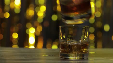 pouring of golden whiskey, cognac or brandy from bottle into glass with ice cubes. shiny background